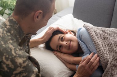 Affectionate military father kissing his daughter who is sleeping on the sofa at home.