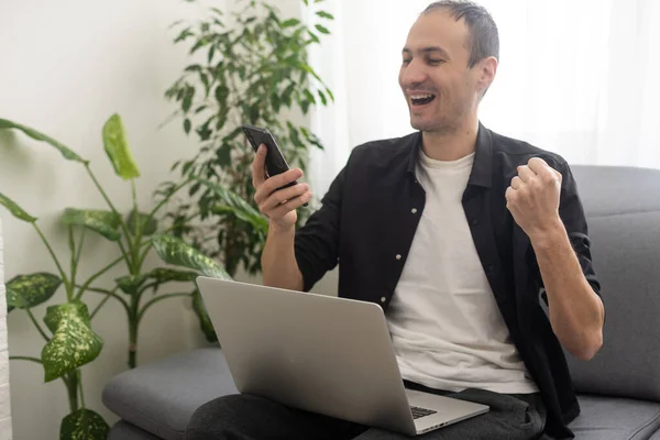Усміхнений Молодий Чоловік Друкує Мобільному Телефоні Сидячи Дивані Вдома Ноутбуком — стокове фото