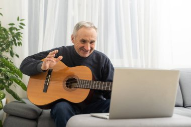 Kulaklıklı, online gitar dersi alan ve laptop ekranına bakan son sınıf öğrencisinin portresi. Emekli erkek, evde bilgisayar başında gitar çalmayı öğreniyor..