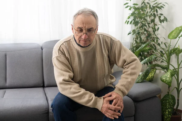 old man with his knee joint pain in sofa, pain in the elderly, health care, elderly care. Elderly man having a knee pain and sitting down. Grandfather with knee pain