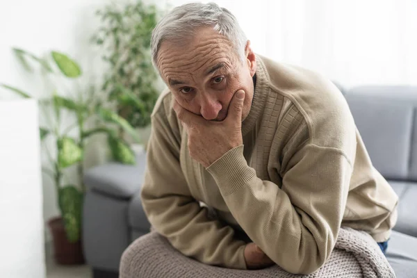Frustrated unhappy middle aged mature man sitting on sofa, feeling depressed alone at home. Confused senior retired grandfather worrying about difficult life decision, copy space, old people solitude.