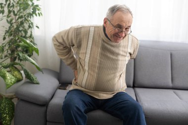Caucasian old senior elderly unhealthy sickness grey male grandpa near sofa at home alone holding hands on back having emergency painful muscle and backache injury problem