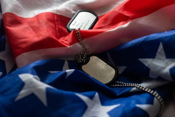 Closeup of a dog tag with the text thank you veterans engraved in it, next to a flag of the United States background