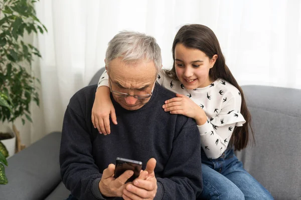 senior grandfather learning to using mobile phone under guidance of pretty young granddaughter sitting on sofa at home.