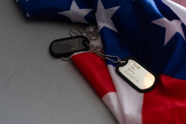 Closeup of a dog tag with the text thank you veterans engraved in it, next to a flag of the United States background