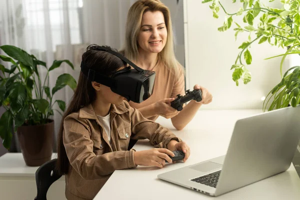 Families and new technologies. Smiling work-at-home mother sitting with daughter wearing vr helmet exploring virtual reality. Curious child teen girl testing AR technology for education.