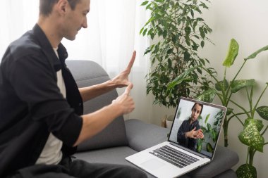 Happy smiling deaf young caucasian man uses sign language while video call using laptop while sitting at home, virtual communication concept