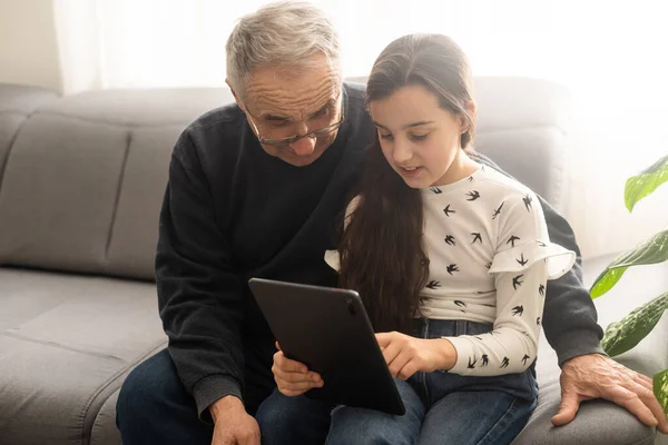 귀여운 소녀와 잘생긴 할아버지는 집에서 소파에 미소짓고 태블릿을 — 스톡 사진
