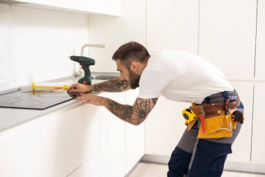 Adam mutfak yüzey dairesini elektrikle tamir ediyor. Tezgaha hob kuruluyor. Adam yeni mutfak mobilyaları ve aletler almış. Elektrikçi ev aletleri kurma işlemi