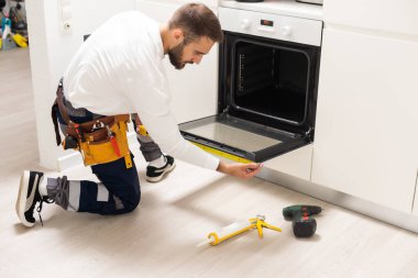 repairman examining oven with screwdriver in kitchen with tool case.