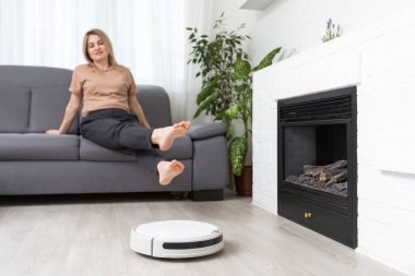 Dinlenen kadının olduğu koltuğun yanındaki modern robot elektrikli süpürge..