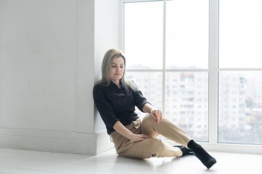 Full length view of young woman near window, wearing black, resting from the business.