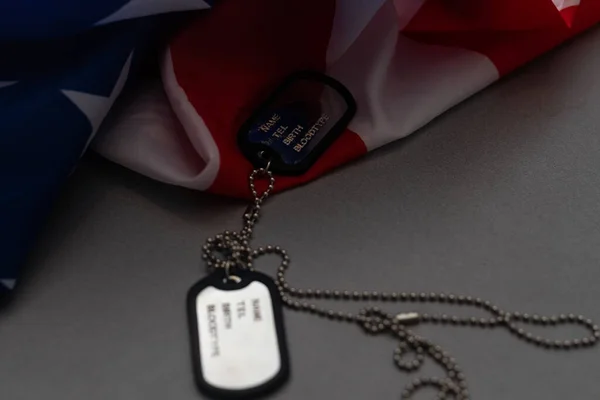 Closeup of a dog tag with the text thank you veterans engraved in it, next to a flag of the United States background