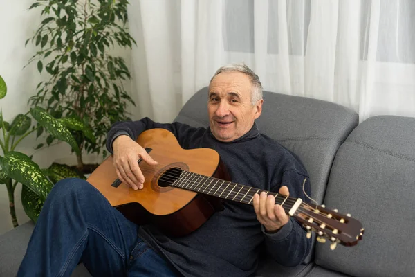 Senior man is playing guitar. Elderly man sitting on the sofa and playing guitar. Portrait of a gray-haired mature man in a sweater learning to play. Enjoying retirement life at home