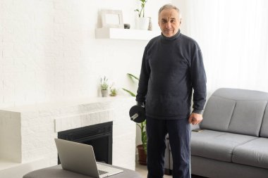 Olgun yaştaki spor. Mutlu orta yaşlı adam evde dizüstü bilgisayarın önünde esneme egzersizleri yapıyor, online dersler izliyor, oturma odasında iç mekanda, boş alanda..