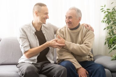 Mutlu iki nesil erkek aile, yaşlı yaşlı yaşlı baba ve gülümseyen yetişkin oğul sohbetin tadını çıkarıyorlar sohbetin tadını çıkarıyorlar arkadaşça olumlu sohbetin tadını çıkarıyorlar evdeki kanepede oturuyorlar.