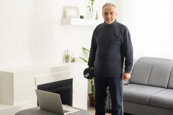 stock image Sport in mature age. Happy middle-aged man doing stretching exercises in front of laptop at home, watching online tutorials, in living room interior, free space.