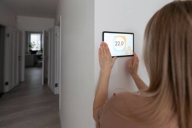 Oturma odasındaki duvara dijital bir dokunmatik ekranla evi kontrol eden kadın..