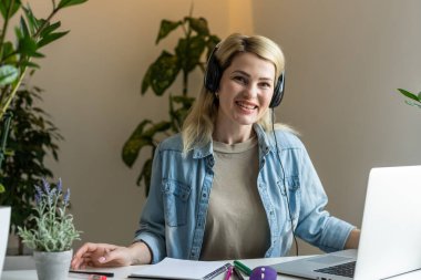 Genç, mutlu, neşeli ve güzel iş kadınının görüntüsü ofiste oturup bilgisayarla kulaklıkla müzik dinliyor..