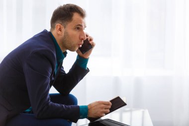 Günlüğünü tutarken kameraya bakan, modern bir şirket ortamında oturan genç ve pozitif bir iş adamının portresi, pencerelerden süzülen hafif güneş ışığıyla.