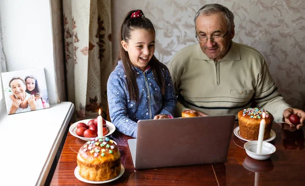 grandfather and granddaughter are talking via video link to their friends. Decorated table with colorful eggs and cake. Chatting during the COVID pandemic and the Easter holidays.