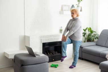 Ev sporu. Aktif kıdemli kadın dizüstü bilgisayarın önünde ısınma egzersizleri yapıyor, çevrimiçi öğretmenlerle eğitim görüyor..