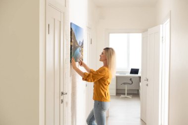Canvas print with gallery wrap interior decor. Woman hangs landscape photography on white wall. Hands holding photo canvas print.