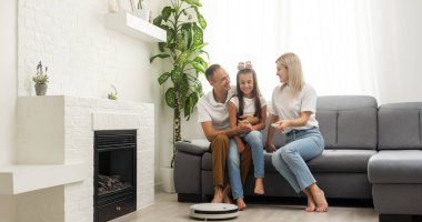 Young family resting on the couch while robotic vacuum cleaner doing its work at home. Household robots concept