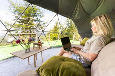 Orta yaşlı bir kadın tatillerde Glamping 'lerin evinde dizüstü bilgisayar kullanıyor. Tatil çadırı. Rahat, kamp, hygge, yaşam tarzı konsepti.