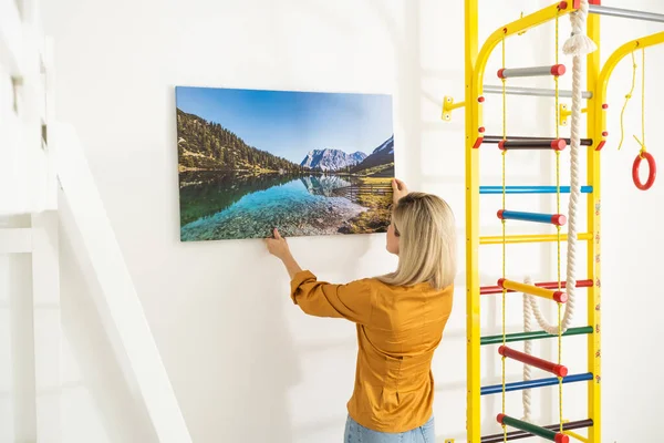 Canvas print with gallery wrap interior decor. Woman hangs landscape photography on white wall. Hands holding photo canvas print.