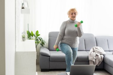 Olgunluk çağında spor. Mutlu son sınıf kadını evde dizüstü bilgisayarın önünde egzersiz yapıyor, online dersler izliyor, boş yer arıyor.