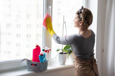 Ev işleri yapan genç bir kadın. Elinde temizlik aletleri olan kadın. Plastik koruyucu sarı eldivenler giyen, elinde bez parçası ve sprey deterjan tutan bir kadın. Bahar temizliği için asla geç değildir..