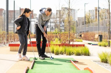 Sharing with golf experience. Cheerful young man teaching his daughter to play mini golf at the day time. Concept of friendly family.