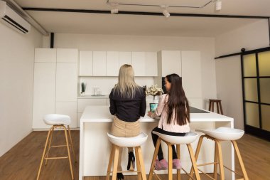 Portrait of mother and daughter sitting in living room. Cute girl holding in her hand a digital tablet while her mom using laptop and looking at something together