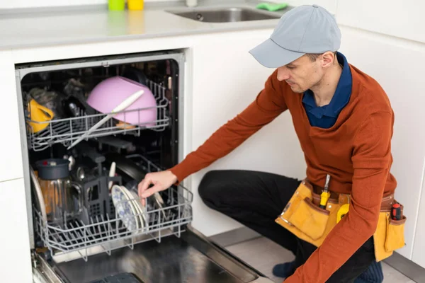 A man or a service worker in special clothes using a screwdriver attaches to the countertop or removes the dishwasher.