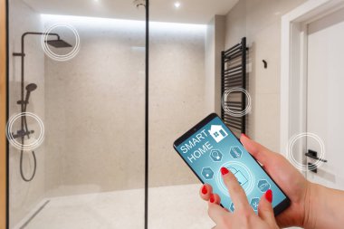 Elinde akıllı telefon tutan ve evinde güvenlik uygulaması olan genç bir kadın. Ev güvenliğini mobil bir cihazdan kontrol etme ve yönetme kavramı.