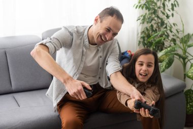 A father and a little daughter play video games using gamepads at home. A man and a child hold game joysticks in their hands. The family is having fun in the bedroom while being quarantined