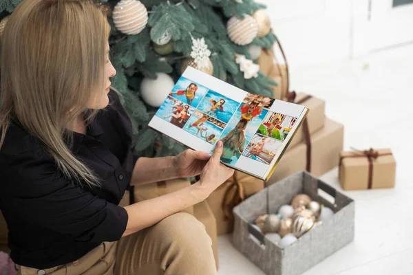 美しく装飾されたクリスマスツリーの近くの床に座っている若い女性は 自宅でクリスマスイブを過ごしながら 古い写真アルバムを見て — ストック写真