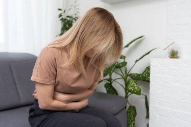 Üzgün, üzgün, mutsuz, karın ağrısı çeken karın ağrısı çeken, gözleri kapalı, regl olan, gıda zehirlenmesi geçiren, ishal olan, oturma odasında kendini rahatsız hisseden bir kadın.