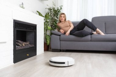 Dinlenen kadının olduğu koltuğun yanındaki modern robot elektrikli süpürge.