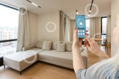 Mobil uygulamada ev sıcaklığı, güvenlik ve çevre kontrolü. Kadın elinde akıllı telefon. Arka planda oturma odası.