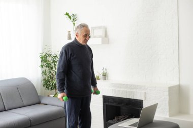 Sport in mature age. Happy middle-aged man doing stretching exercises in front of laptop at home, watching online tutorials, in living room interior, free space.