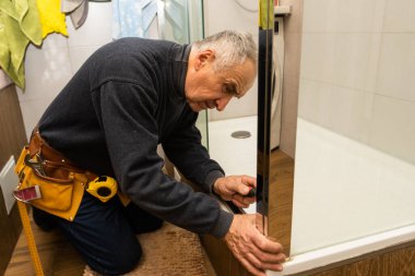 an elderly man repairing door of shower cabin in bathroom.