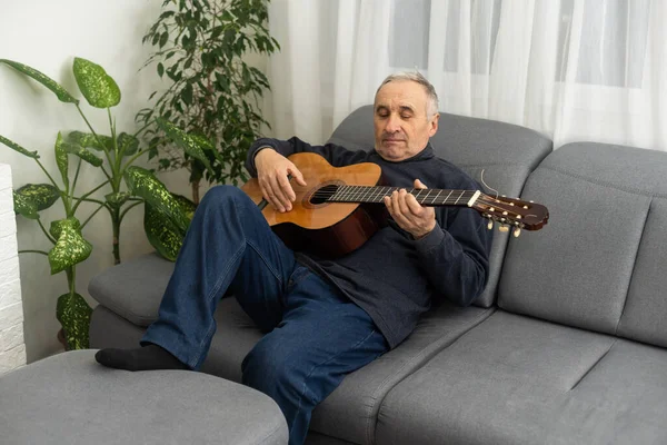 stock image Senior man is playing guitar. Elderly man sitting on the sofa and playing guitar. Portrait of a gray-haired mature man in a sweater learning to play. Enjoying retirement life at home