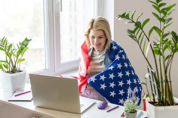 stock image Happy woman employee sitting wrapped in USA flag, shouting for joy in office workplace, celebrating labor day or US Independence day. Indoor studio studio shot isolated on yellow background.