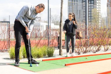 Sharing with golf experience. Cheerful young man teaching his daughter to play mini golf at the day time. Concept of friendly family.