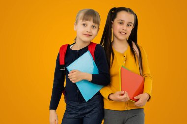 Education Concept. Set portrait of diverse schoolchildren holding stack of academic books and wearing backpack, yellow orange color studio background.