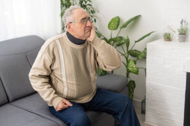 Diş ağrısı çeken, yanağa dokunan yaşlı bir adam koltukta oturduğu için acı çekiyor.