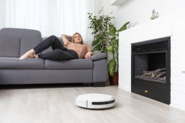 Dinlenen kadının olduğu koltuğun yanındaki modern robot elektrikli süpürge.
