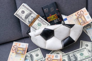 Çevrimiçi spor bahsi. Akıllı telefon ve futbol topu olan bir elin arka planına dolar düşüyor. Yaratıcı altyapı, kumar